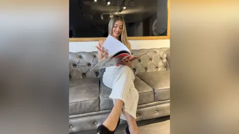 Smiling woman in a white knit maxi dress reading a book on a grey tufted sofa in a bright studio.