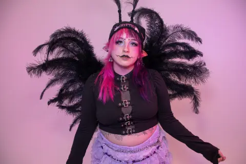 Woman with pink hair wearing black fairy wings and a gothic crop top against a pink background.