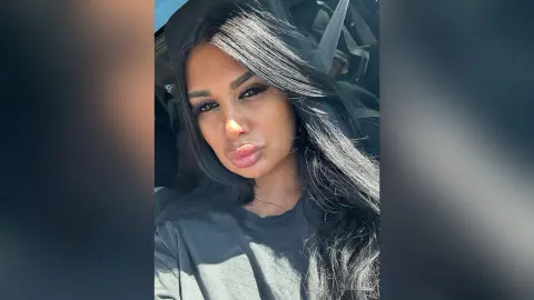 Brunette woman in a black t-shirt sitting in a car with long flowing hair and a relaxed expression.