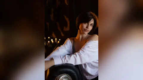 Elegant woman in a white off-the-shoulder shirt posing in a moody, dimly lit room with candlelight.