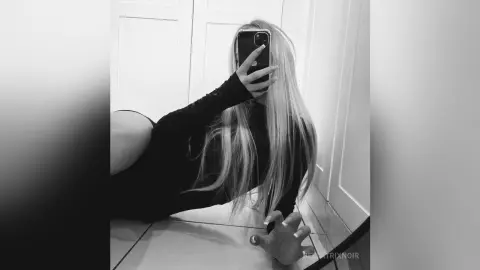 Blonde woman in a black long-sleeve top taking a moody black and white mirror selfie in a modern room.