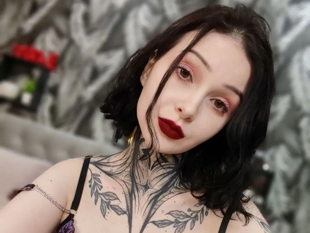 Young woman with dark hair and floral chest tattoos wearing a black strap top in a modern indoor setting.