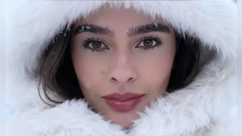 Close-up portrait of a woman with natural makeup wearing a cozy white faux fur hooded coat in a snowy setting.