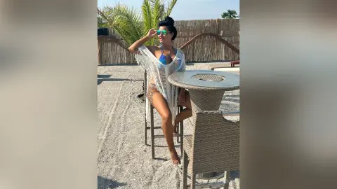 MissyJolie sits on a tall stool outdoors wearing a blue bikini and a white fringed cover-up near a woven table.