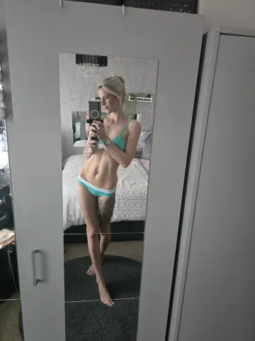Blonde woman in mint green bikini posing for a mirror selfie in a modern bedroom.