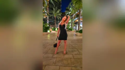 Glamorous woman in a black cocktail dress and gold heels posing on a city walkway at night.
