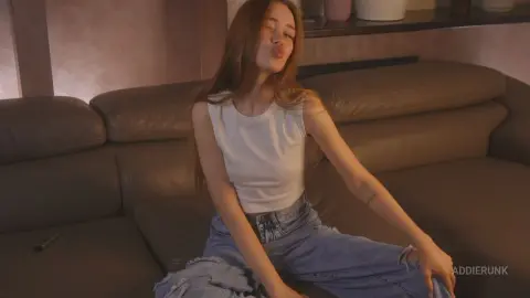 AddieRunk wearing a white tank top and ripped blue jeans while sitting on a brown leather couch.