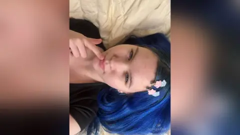 DeidreSarin lying on a white bed with her blue hair and a finger touching her lip.