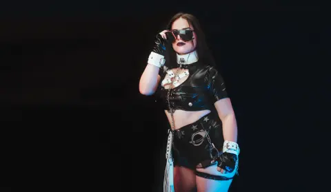 AbbyShinee wears a black shiny crop top and matching shorts with white trim, posing against a dark background while wearing large black sunglasses.