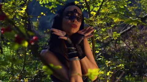Mysterious woman in a black lace mask and fishnet gloves posing amidst lush green forest foliage.