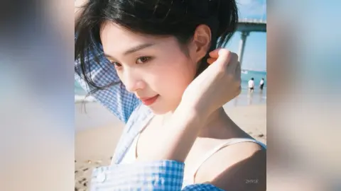 ZhiYue poses on a sunny beach wearing a white tank top and a blue checkered shirt, running her hand through her dark hair.