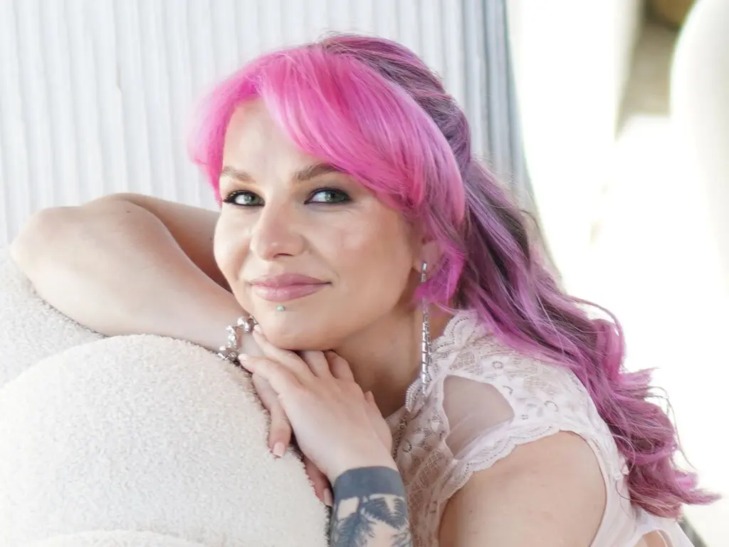 Smiling woman with bright pink hair wearing a white lace top in a bright, airy setting.