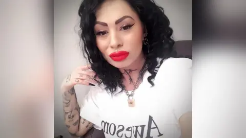 DeeaAnderson poses with long wavy black hair and bright red lipstick, wearing a white t-shirt and a silver chain necklace.