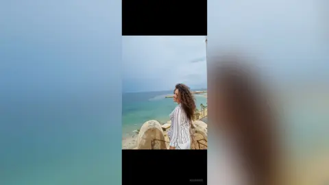 Dreamy portrait of a woman in a white striped shirt overlooking the turquoise ocean at a scenic coastal viewpoint.