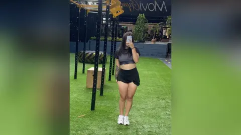 NatiSweet stands outside in a black crop top and short black skirt, posing for a mirror selfie on green grass.