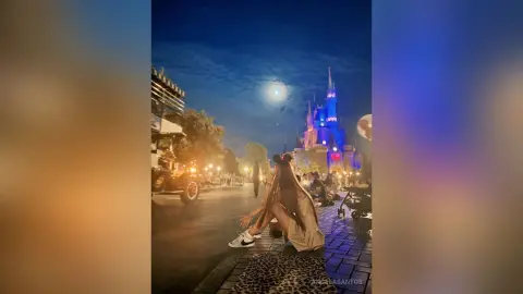 Young woman in casual outfit sitting on a curb at night near Disney World castle under a bright moon.