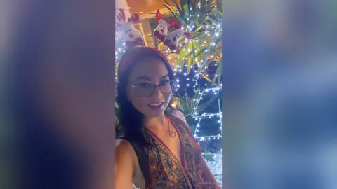 Smiling woman in glasses and floral dress posing in front of festive Christmas lights and holiday decor.