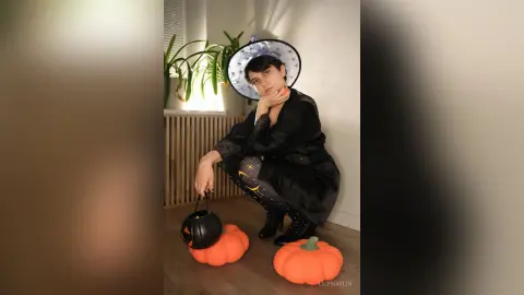 Woman in a black witch costume and hat holding a pumpkin bucket in a cozy indoor Halloween setting.