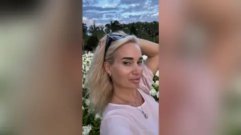 Smiling blonde woman in a pink t-shirt relaxing outdoors in a lush garden under a cloudy sky.