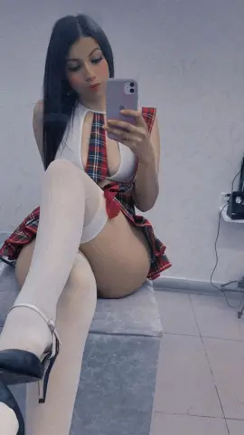 Young woman in a plaid schoolgirl outfit and white tights posing for a selfie in a modern room.