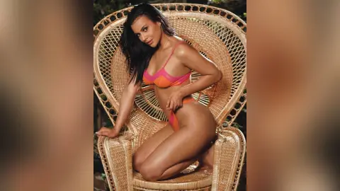 Brunette woman in an orange bikini sitting on a vintage wicker chair in a warm, tropical setting.