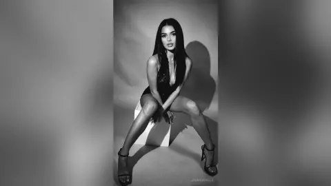 JasmineHille poses sitting on a white block wearing a black bodysuit and high-heeled sandals against a plain background.