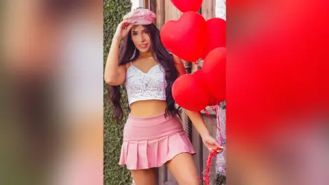 Smiling woman in white lace crop top and pink skirt holding red heart balloons outdoors.