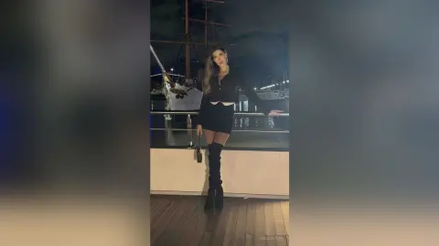 Stylish woman in a black mini dress and over-the-knee boots posing on a wooden pier at night by the water.
