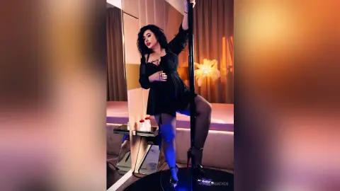 AdiraMaeven poses with a dance pole, wearing a black sheer babydoll outfit and black stockings with high heels.