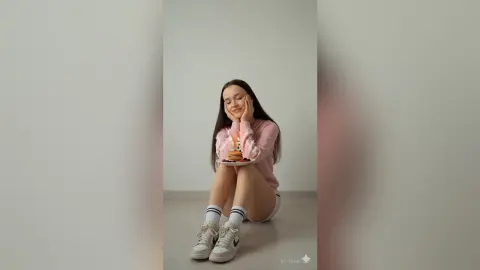 Happy young woman in pink sweater sitting on floor holding a small birthday cake in a minimalist room.