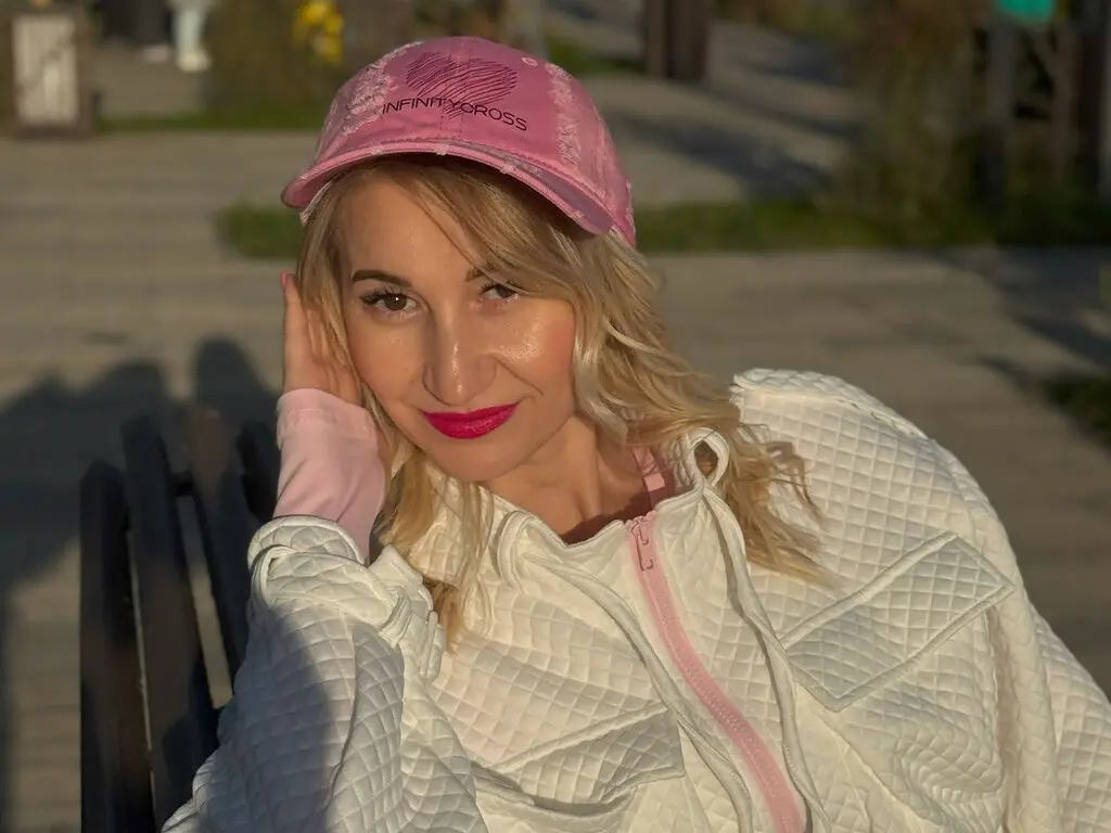 CandiceBright wearing a pink cap and white quilted jacket, posing outdoors on a bench with her hand against her face.