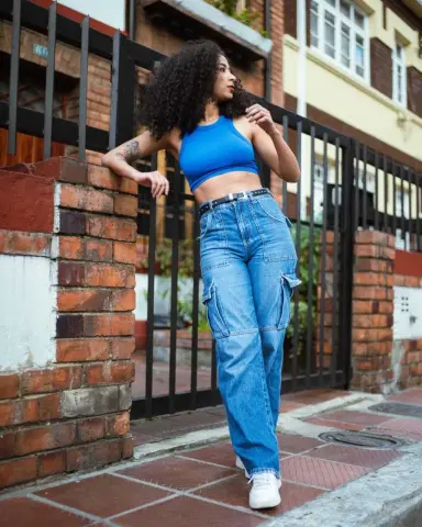 Young woman in blue crop top and baggy denim jeans leaning against a brick wall in a sunny urban setting.