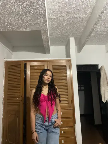 Young woman with long curly hair wearing a pink halter top and blue jeans in a cozy indoor setting.