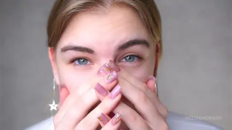 Young woman with long manicured nails covering her face against a neutral grey background.