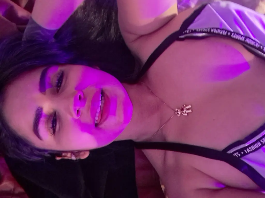 lilith_zaffron smiles while posing with a peace sign, showing her long black hair and wearing a black top.