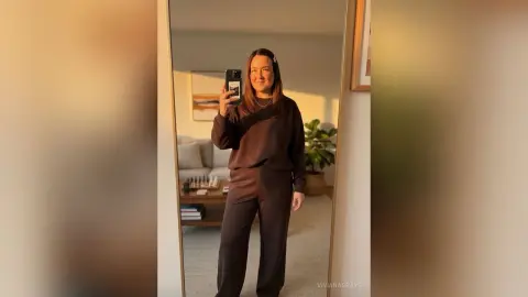 Woman in a cozy dark brown lounge set taking a mirror selfie in a bright, modern living room.