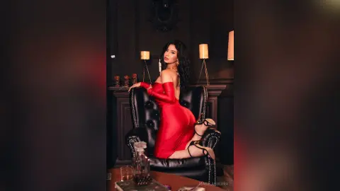 Elegant woman in a red satin dress sitting on a black leather chair in a moody, dark luxury interior.