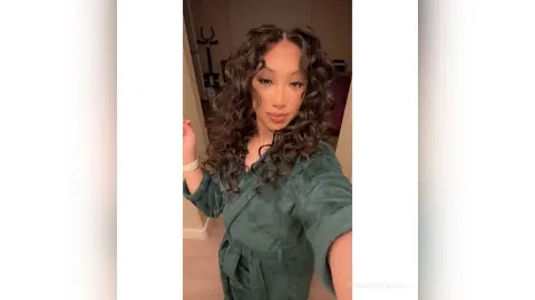 Smiling woman with long curly hair wearing a cozy dark green velvet robe in a bright room.