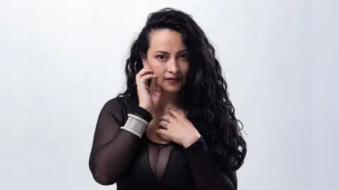 Portrait of a woman with long curly dark hair wearing a black sheer top against a white background.