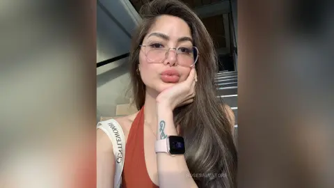 Young woman with glasses and an orange top posing thoughtfully in a modern sunlit interior.