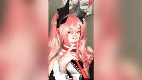 Cosplayer with long pink hair and black devil horns wearing a gothic white and black outfit in a soft pink setting.