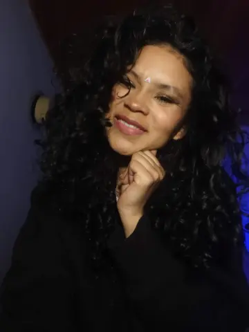 Hathorlove smiles with her hand under her chin, showing off her long, dark curly hair and a small white triangle mark on her forehead.
