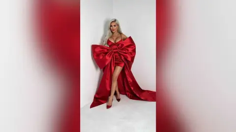 Glamorous blonde woman in a stunning red satin gown with a massive bow detail posing in a studio.