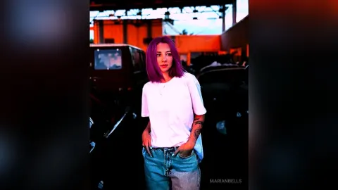 MarianBells — MarianBells with purple hair wears a white t-shirt and blue jeans while standing in a garage.