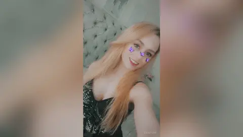 Blonde woman in a black floral tank top smiling in a bright, cozy bedroom setting.