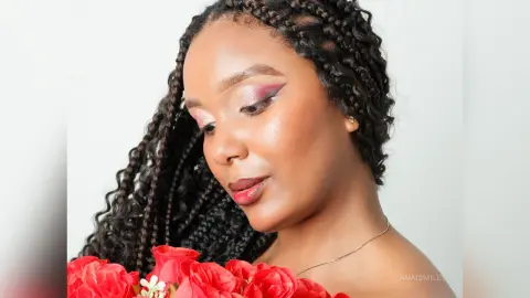 Woman with long dark braids and glowing makeup holding a bouquet of red roses against a white background.