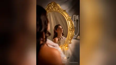 Elegant woman in a white lace dress looking into a vintage gold mirror by candlelight in a moody setting.