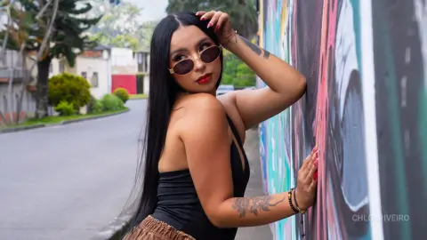 ChloeRiveiro — Stylish woman in a black bodysuit and sunglasses leaning against a colorful graffiti wall on a city street.