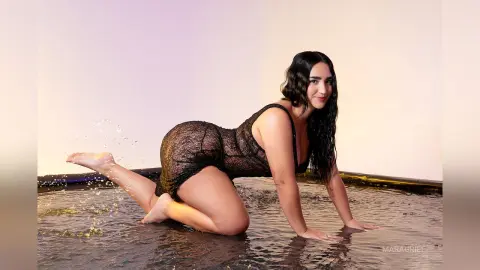 MaraGrief poses on all fours in a shallow pool of water, wearing a black lace bodysuit that shows her curves and wet black hair.