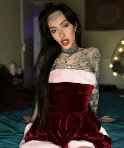 Tattooed woman in a red velvet Santa outfit sitting on a bed with a moody, festive atmosphere.